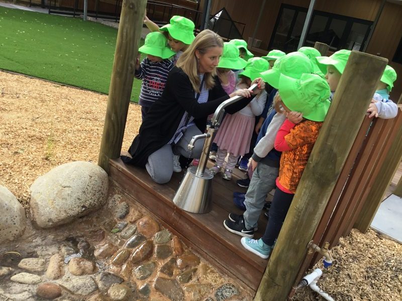 An adult kneels beside four young children on a wooden bridge as they all use a handwashing station together outdoors, embracing a philosophy of learning through shared experience.