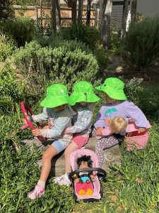 An adult and 3rd old group wearing bright hats are playing outside on artificial grass, with three children sitting on a blue play structure shaped like a boat.