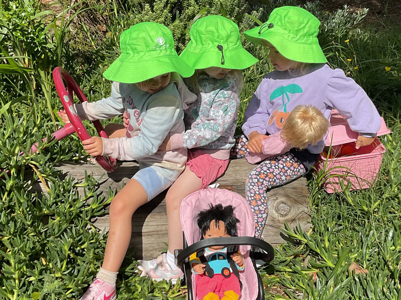 Three young children wearing bright green hats sit on a log outdoors, pretending to drive with a steering wheel, with two dolls placed in front of them.