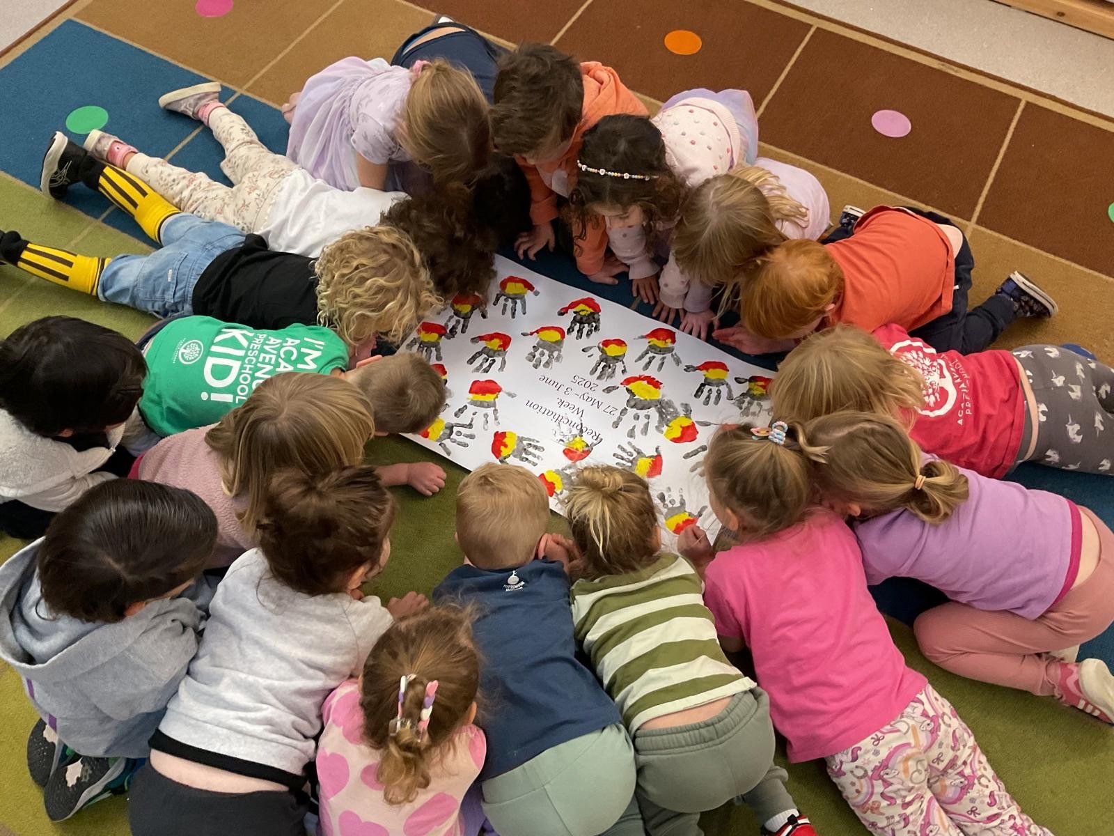 A group of young children lies in a circle on a colorful rug, closely looking at a large sheet of paper with handprint artwork and writing at the center.