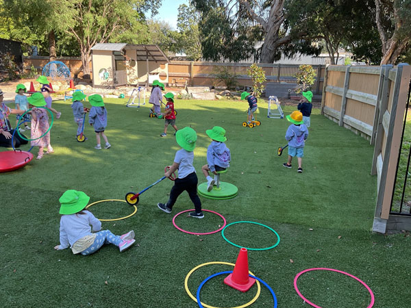 Children wearing green hats play with hoops and toys on a grassy outdoor playground, surrounded by trees and a wooden fence, exploring the simple philosophy of joy through play.
