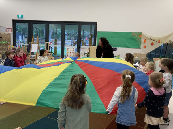 A group of children and an adult hold the edges of a colorful parachute in a classroom, participating in an activity together.