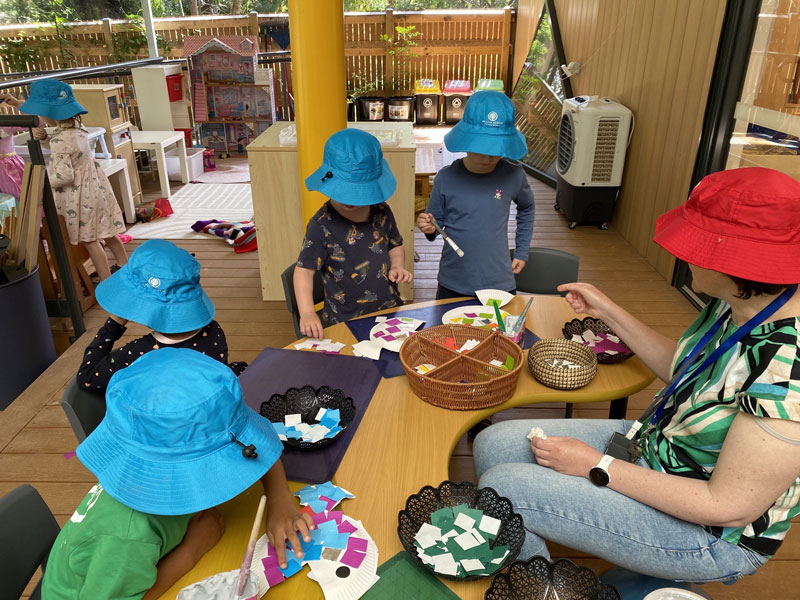 Three young children wearing bright green hats sit on a log outdoors, pretending to drive with a steering wheel, with two dolls placed in front of them.