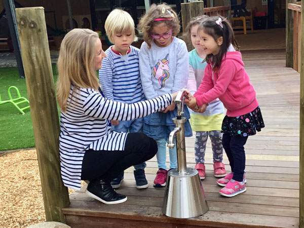 An adult kneels beside four young children on a wooden bridge as they all use a handwashing station together outdoors, embracing a philosophy of learning through shared experience.