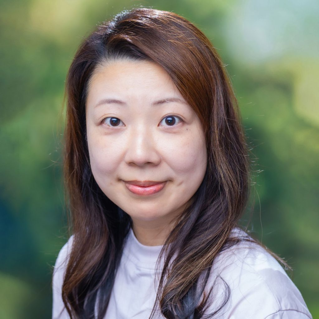 A woman with long dark hair and a light top is looking at the camera, with a blurred green background behind her.