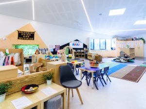 A brightly lit preschool classroom designed for play, featuring various activity stations, small tables and chairs, shelves with toys, and colorful rugs on the floor.