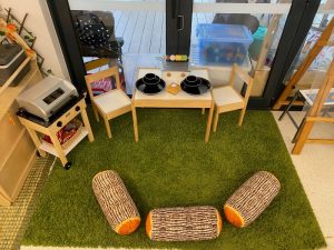 Children’s play kitchen setup with a small table, two chairs, toy stove, and three log-shaped cushions on a green rug invites creative play in front of large glass doors.