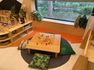 A low wooden play table with colorful interlocking sticks sits on a round rug, surrounded by green-patterned floor cushions in a bright room with shelves, toys, and a large window.