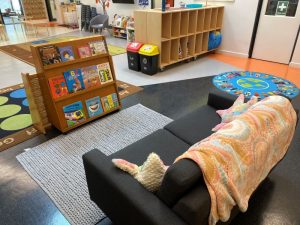 A children's reading area designed for play, featuring a gray couch, colorful blanket, bookshelf of picture books, cubbies, rugs, and two recycling bins in a vibrant classroom setting.
