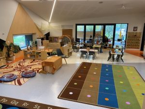 A brightly lit preschool classroom with colorful rugs for play, small tables and chairs, engaging learning materials, and large windows overlooking an outdoor area.