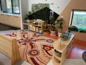 A cheerful children's playroom with a patterned rug, low wooden shelves, toys for play, books, potted plants, and a cozy window seat with cushions.