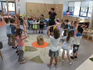 A group of children play in a circle, raising wooden sticks and following an adult instructor in a brightly lit classroom.