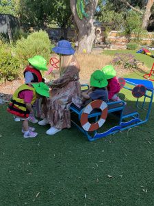 A woman in a sunhat sits on a bench outdoors surrounded by four young children in green hats, with two sitting on a blue play structure shaped like a vehicle.