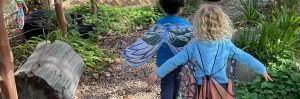 Two children wearing butterfly wings walk along a gravel path in a garden with greenery and logs on either side.