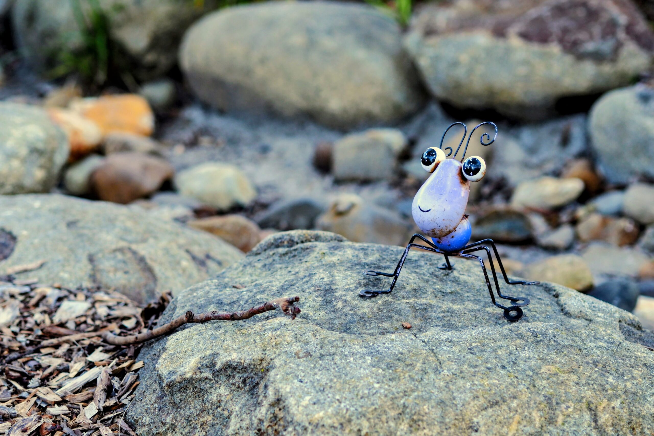 A small, whimsical metal insect sculpture with big eyes sits on a rock among natural stones and dirt outdoors.
