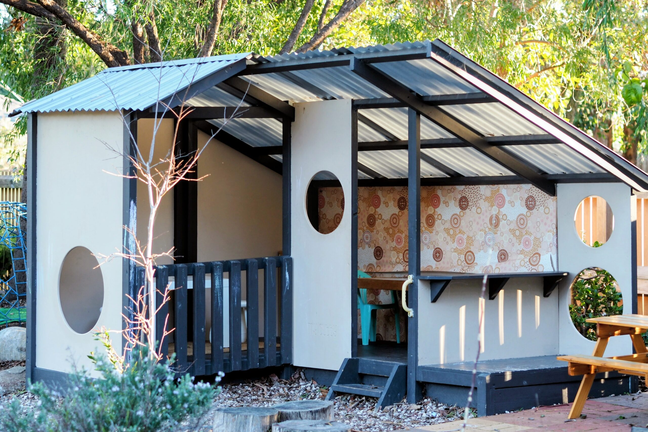 A small modern playhouse with a metal slanted roof, circular windows, a small porch, and patterned wallpaper inside, set in an outdoor area with trees and a picnic table nearby.