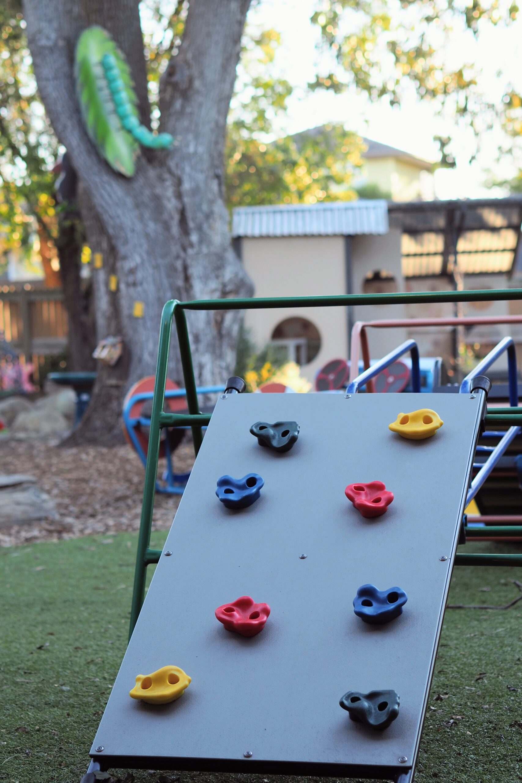 Children’s climbing wall with colorful handholds in an outdoor playground, with trees and playground equipment in the background.