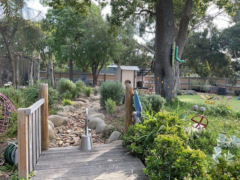 A wooden footbridge leads over rocks to a garden with trees, bushes, a playhouse, and various outdoor decorations, under a clear sky.